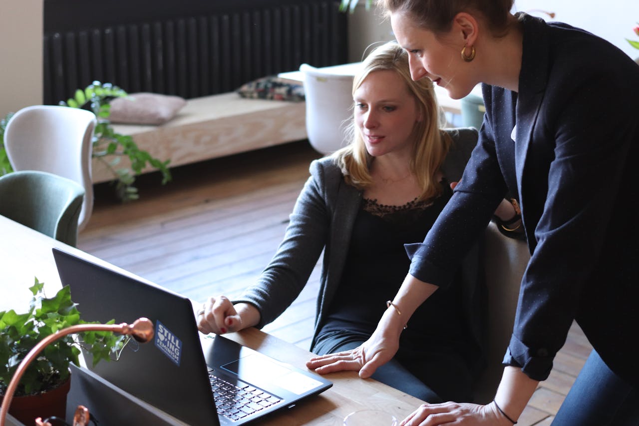 hero-img Two professional women discuss a project using a laptop in a modern office environment.